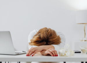 Donna seduta alla scrivania con la testa appoggiata sulle braccia, visibilmente stanca, accanto a un laptop e fazzoletti.