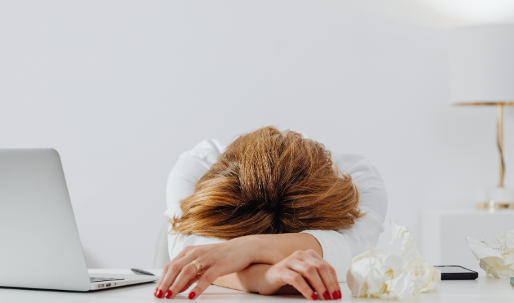 Donna seduta alla scrivania con la testa appoggiata sulle braccia, visibilmente stanca, accanto a un laptop e fazzoletti.