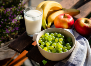 Ciotola di uva verde con bicchiere di latte, mele e banane su tavolo all’aperto