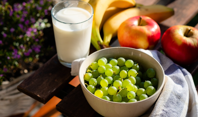 Ciotola di uva verde con bicchiere di latte, mele e banane su tavolo all’aperto