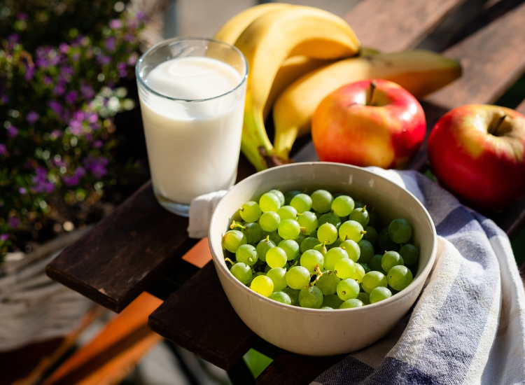 Ciotola di uva verde con bicchiere di latte, mele e banane su tavolo all’aperto